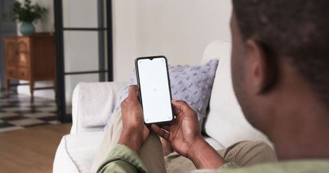 African american man sitting on sofa holding smartphone with blank screen for app mockup