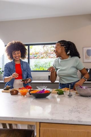 African american friends laughing while cooking at marble kitchen island using smartphone