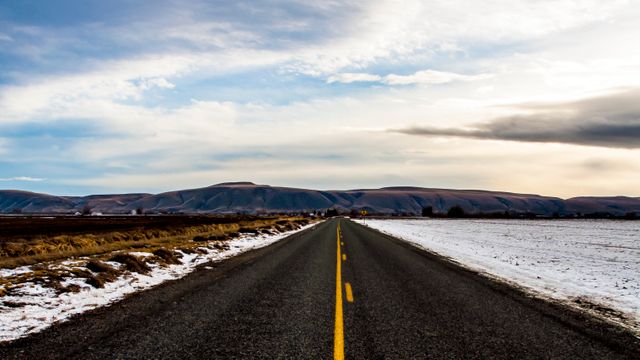 The image features an open road extending through a snow-covered landscape with distant mountains, under a dramatic sunset sky. Ideal for themes related to travel, journey, adventure, exploring nature, and winter scenery. It can be used for blog headers, travel websites, inspirational posters, and advertisements for road trips or winter getaways.
