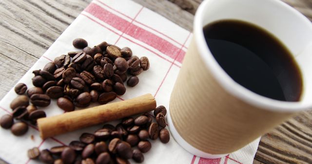 Cup of black coffee in paper cup placed on wooden table with coffee beans and cinnamon stick next to it on checkered napkin. Perfect for use in articles on coffee culture, morning routines, advertising coffee-related products, or evoking a rustic, cozy atmosphere in social media posts.