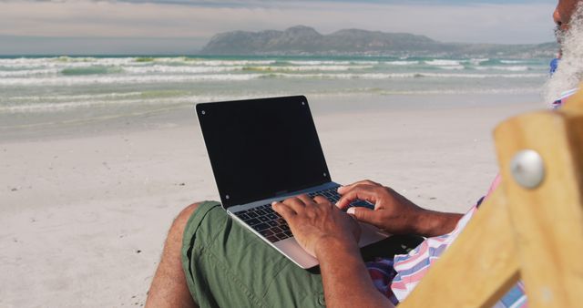 View of senior man using laptop while sitting in a folding chair on a sunny beach. He is focused on his work while enjoying beautiful coastal scenery. Ideal for concepts like remote work, retirement lifestyle, freelancing, work-life balance, and tropical vacations.