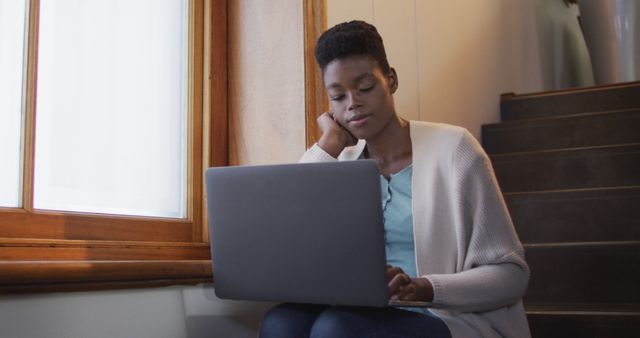 Thoughtful african american attractive woman sitting on stairs and using laptop. leisure time, wellbeing and relaxing at home.