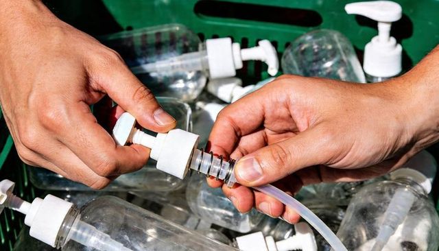 Adult hands assembling soap pump dispenser spring and tube on workbench with empty bottle crate