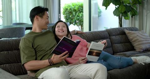 Asian couple reading on tufted gray sectional sofa in sunlit living room, cozy romance