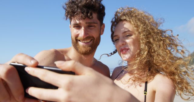 Young couple taking a selfie on a sunny beach. They look happy and relaxed, enjoying their vacation by the sea. Ideal for ads and promotions related to travel, vacations, summer activities, and tourism. Can be used to showcase happy moments, youth culture, and leisure activities.