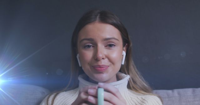 Young woman enjoying a pleasant moment at home, holding a coffee mug and wearing wireless earbuds. Perfect for promoting lifestyle blogs, coffee brands, technology gadgets, and home comfort. This shot captures a cozy and modern vibe suitable for advertising relaxation or contemporary living.