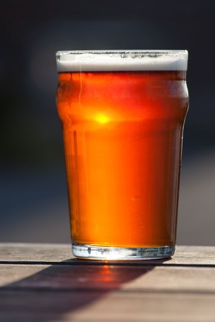 A frothy pint of amber beer sits on an outdoor table, with golden sunlight illuminating the drink. Ideal for use in advertisements for breweries, pubs, summer events, outdoor dining, or lifestyle blogs focusing on beverages and relaxation.