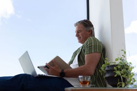 Mature man writing in notebook while using laptop on sunlit window seat home workspace