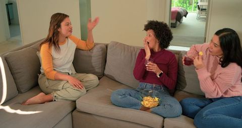 Diverse friends laughing, sharing chips and red drinks on modern gray l-shaped sofa
