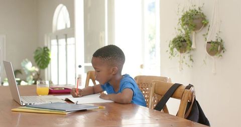 African american boy doing homework at home with laptop and notebook in bright dining room