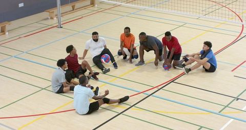 Multiracial male volleyball team sitting in circle on indoor court practicing teamwork