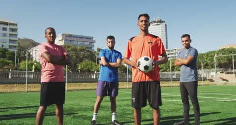 A group of diverse male soccer players in bright sportswear confidently standing on a green playing field under a clear sky. One player in the middle foreground is prominently holding a soccer ball. This inclusive team includes a disabled athlete, enhancing themes of diversity and determination. Ideal for content promoting sportsmanship, inclusivity, team building in urban environments, or inspirational stories about overcoming challenges.