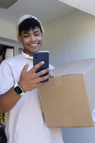 African american man smiling while holding delivery box and checking smartphone at doorstep
