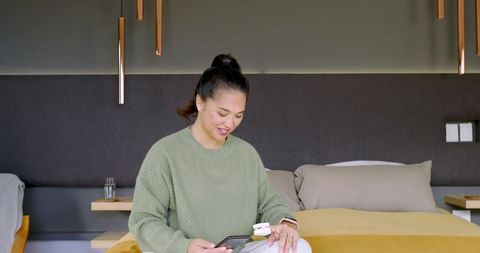 Asian woman using pulse oximeter while relaxing with smartphone on cozy bed