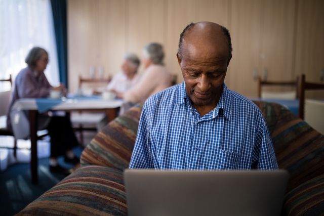 Senior man sitting on sofa using laptop in a nursing home. Ideal for illustrating concepts of elderly people embracing technology, senior living, and digital literacy among older adults. Can be used in articles, advertisements, and brochures related to senior care, retirement homes, and technology for seniors.