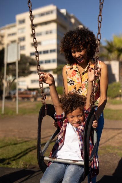 Front view of a biracial woman and her son enjoying free time together at a playground on a sunny day, the boy sitting on a swing while his mother pushes him, both of them smiling