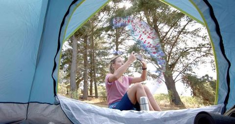 Woman sitting in tent holding smartphone as iridescent ribbon streams across sunlit forest