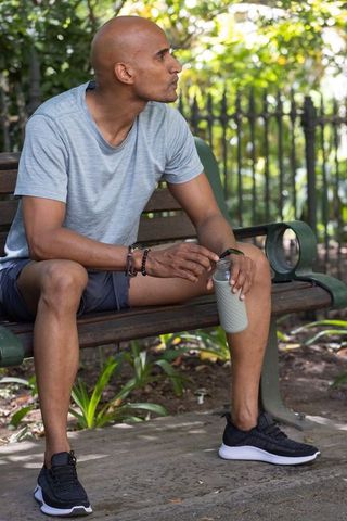 African american man resting on park bench holding reusable water bottle in leafy park