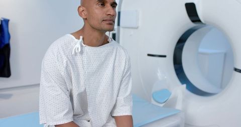 African american man sitting by ct scanner wearing hospital gown preparing for ct scan