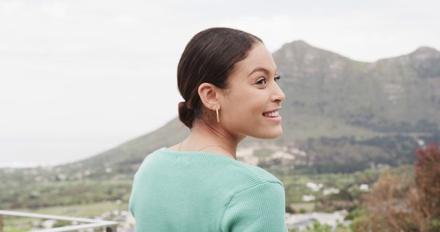 Young woman smiling and enjoying a scenic mountain view. Surrounded by lush greenery, she appears calm and content. Ideal for advertisements, lifestyle blogs, travel websites, promotional materials for holiday packages, or mental wellness campaigns.