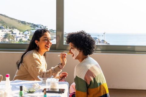 Hispanic and african american women applying facial mask by oceanview window for selfcare