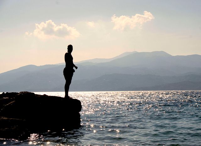 Person is standing on a rocky cliff with their silhouette highlighted against the sparkling sea and a mountainous backdrop during sunset. This evokes a serene and contemplative mood, perfect for use in travel brochures, motivational posters, or relaxation content. The tranquil and scenic atmosphere also makes it suitable for nature documentaries or landscape photography collections.