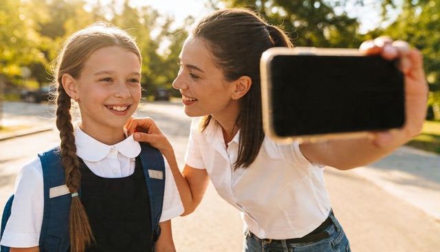 Mother adjusting daughter's school uniform while taking morning selfie on sunny sidewalk