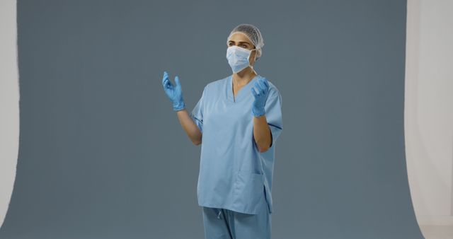 Female healthcare worker standing in blue scrubs, wearing a mask, surgical gloves, and a hair net against a solid blue background. Ideal for illustrating concepts related to healthcare industry, medical professions, safety, patient care, and hospital environments.