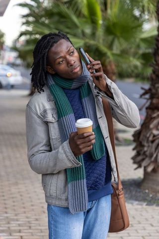 Talking on phone holding coffee on palm-lined sidewalk wearing denim jacket and scarf