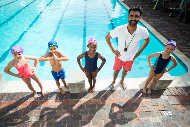 Male swim instructor standing with children at poolside, all wearing swimwear and goggles. Ideal for use in materials promoting swimming lessons, water safety, summer activities, and children's fitness programs.
