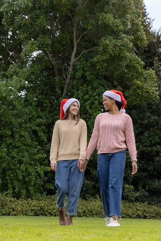 Diverse women walking hand-in-hand wearing santa hats and cozy sweaters and jeans in park