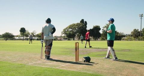 African american batsman facing bowler in park cricket match, teammates fielding