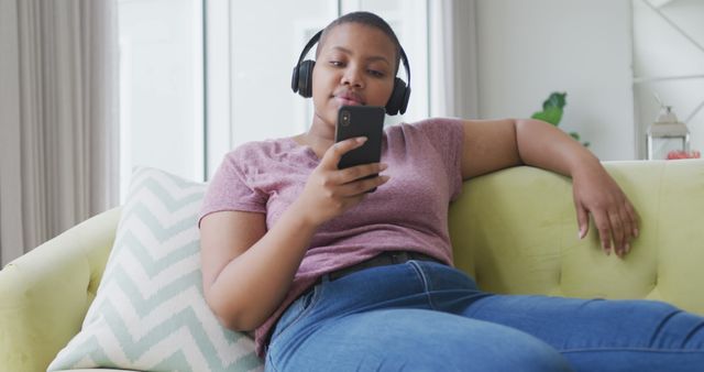 Young adult lounging on a couch, wearing headphones and using a smartphone. Suitable for themes of relaxation, leisure, technology usage, casual home life, listening to music, and digital lifestyles.