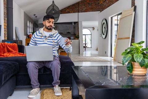 Asian man in 30s working on laptop and holding mug in modern open-plan living room