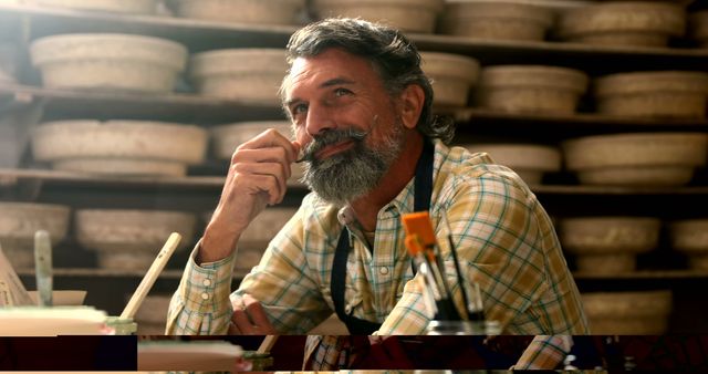 A middle-aged Caucasian man with a beard smiles thoughtfully in a workshop, surrounded by shelves of what appear to be cheese wheels, with copy space. His expression and the artisanal setting suggest he may be a craftsman or cheesemaker taking pride in his work.