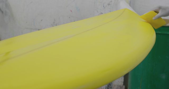 Person sanding yellow surfboard in workshop, working on detail of board's surface. Ideal for use in articles about surfboard manufacturing, craftsmanship, and custom surfboard creation.
