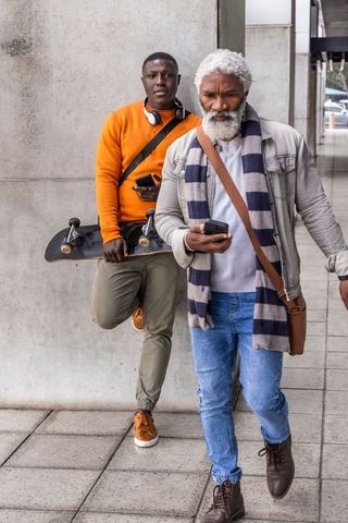 Two men walking urban sidewalk, one checking phone while other leaning with skateboard