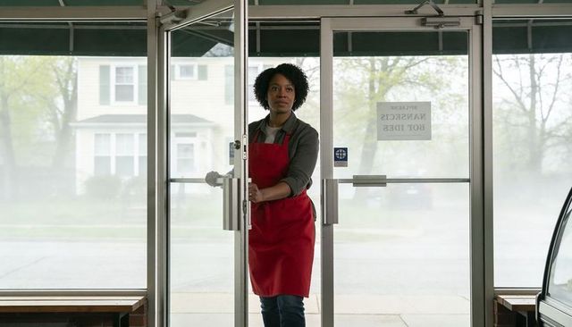 Black shop employee opening glass door wearing red apron at quiet neighborhood storefront