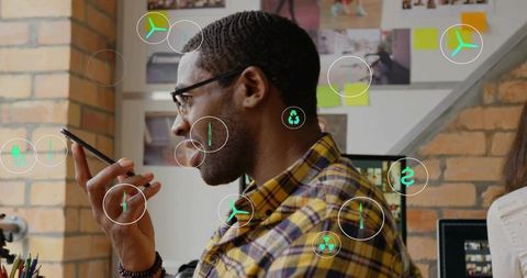 Creative professional speaking into smartphone at office desk with tech icons overlay