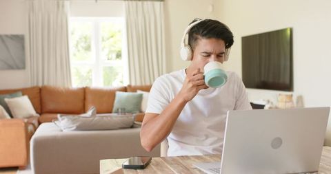 Young man sipping mint mug while working on laptop at home wearing headphones in cozy living room