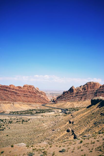 Winding road cutting through rugged canyon landscape under bright blue sky, showcasing dramatic rock formations and dry, arid terrain. Ideal for travel and adventure themes, desert landscapes, road trip promotions, and nature photography blogs.