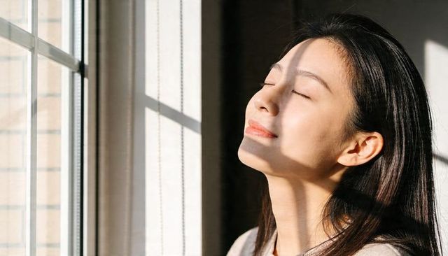 Asian woman basking in morning sunlight by window with serene smile and closed eyes