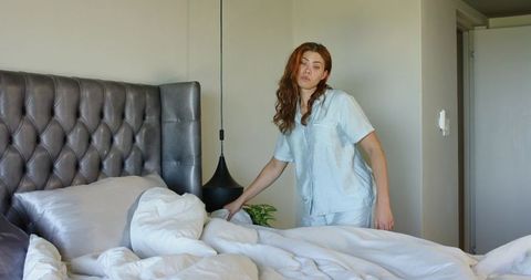 Woman making bed in light blue pajamas smoothing duvet and pillows in minimalist bedroom