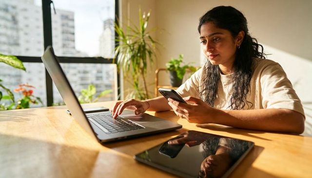 Sunlit indian woman working from home, typing on laptop and checking phone in urban apartment
