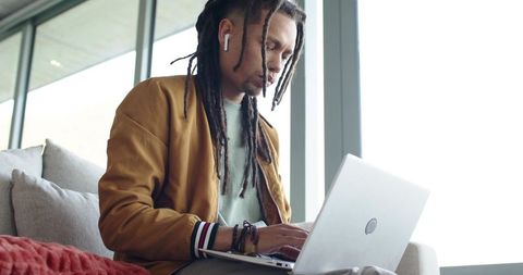 Young man with dreadlocks focusing on laptop in sunlit modern lounge wearing earbuds