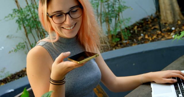 Young woman with pink hair and glasses seated outdoors, using smartphone and laptop. She is smiling while holding the phone near her mouth, suggesting she is using a voice feature. This can be used to illustrate themes like technology use, outdoor working, communication, and modern lifestyle.