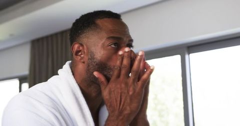 Mature african man grooming face in white bathrobe near bright window, serene skincare
