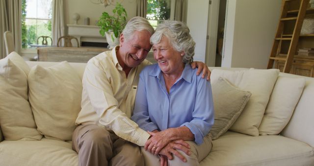 Elderly couple sitting and laughing together on a couch in a cozy living room. Ideal for content focusing on family, love, aging, happiness, and home life. Can be used in articles, blogs, or advertisements related to senior living, retirement, or home decor.