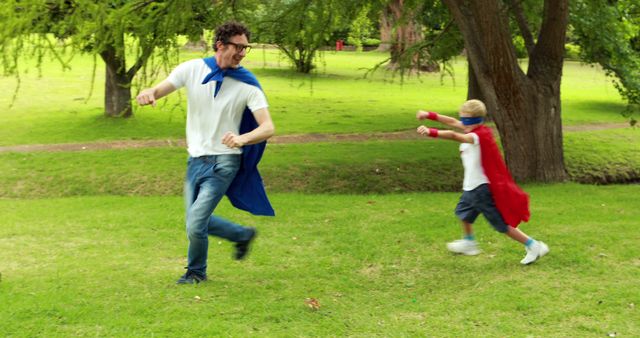 Father and young son dressed as superheroes are playing and running on a sunny day at a park. Both are wearing capes and are enjoying imaginative play together. This can be used for topics related to family bonding, childhood activities, outdoor play, parenting tips, or seasonal recreation.