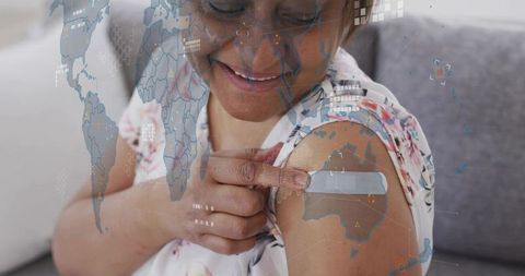Smiling senior woman pointing to vaccine bandage on arm with world map and data overlay